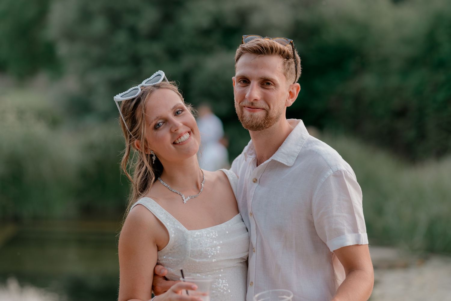 Smiling couple in white outfits posing together outdoors near a sunlit riverbank.