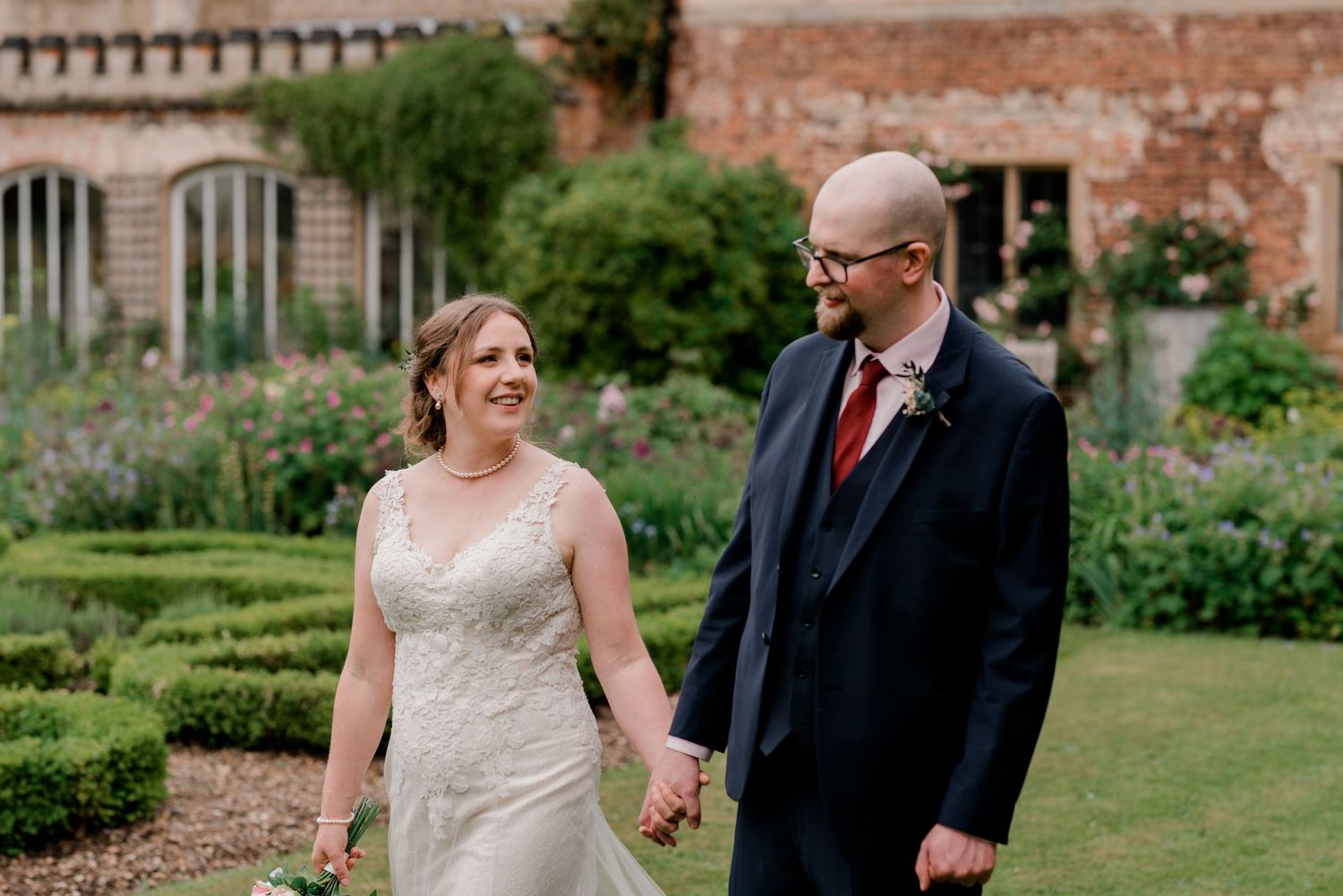 Smiling bride and groom in navy suit stroll hand in hand through elegant formal garden.