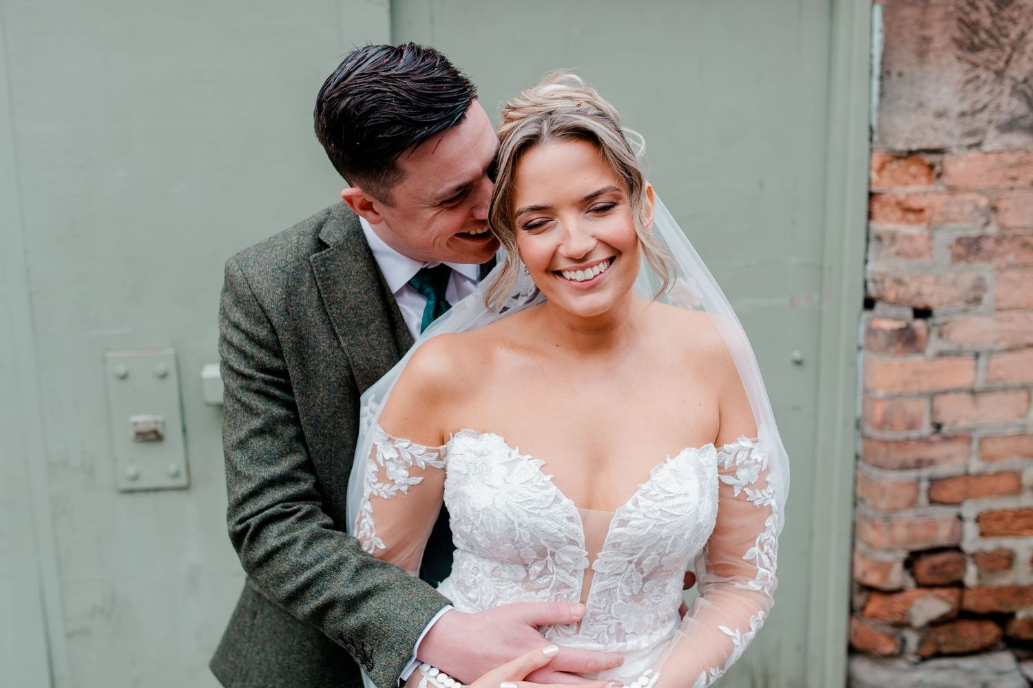 Groom whispers to smiling bride in lace long-sleeve gown against a rustic brick wall.