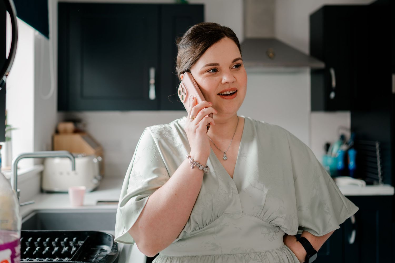 Woman in sage green wrap dress talking on phone in modern kitchen with dark cabinets.