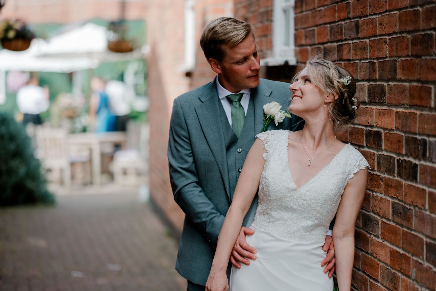 Bride and groom share a romantic moment leaning against a brick wall, groom in gray suit, bride in white lace dress.