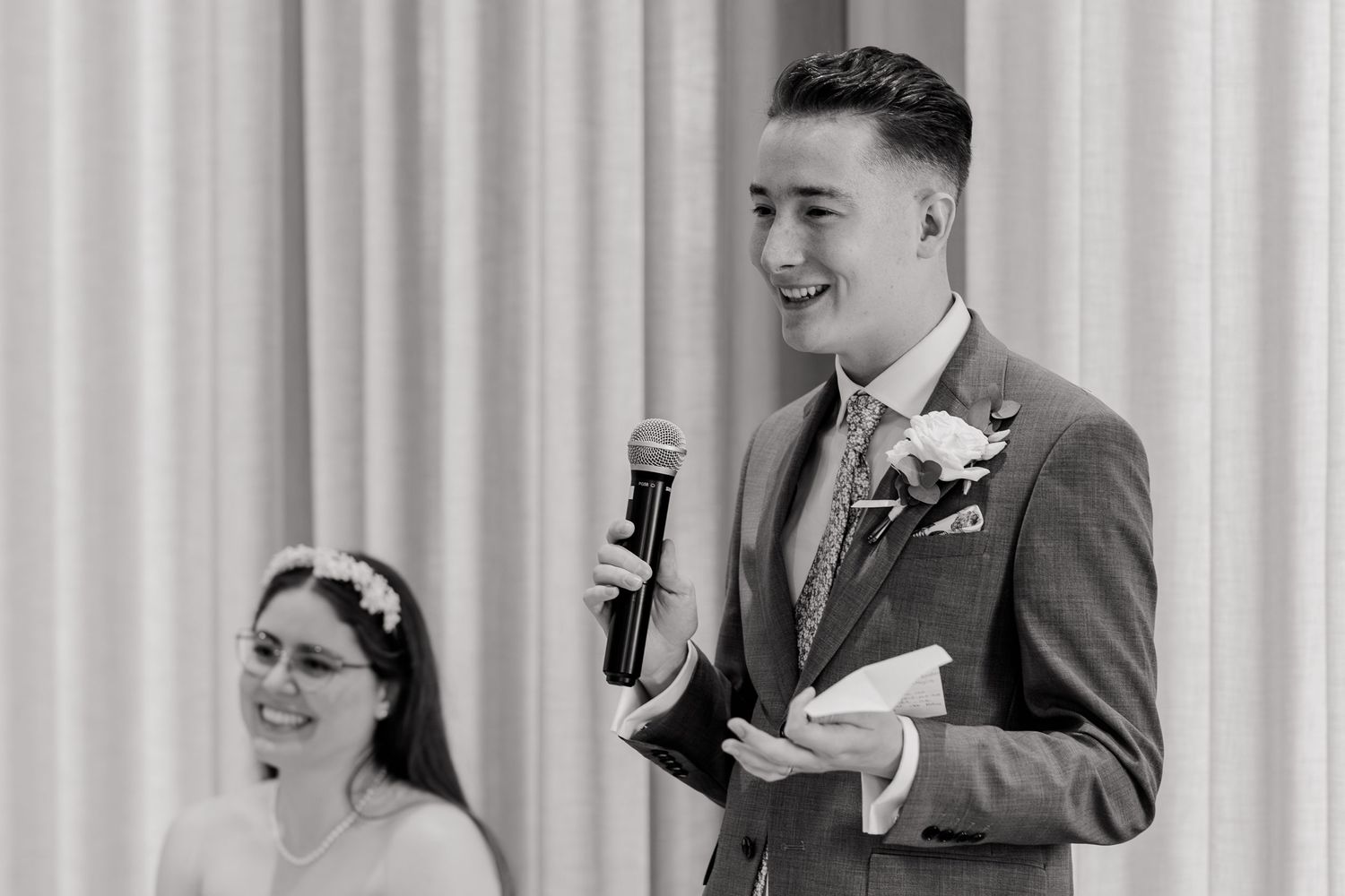 Groom in gray suit speaking into microphone at wedding reception while bride laughs in background.