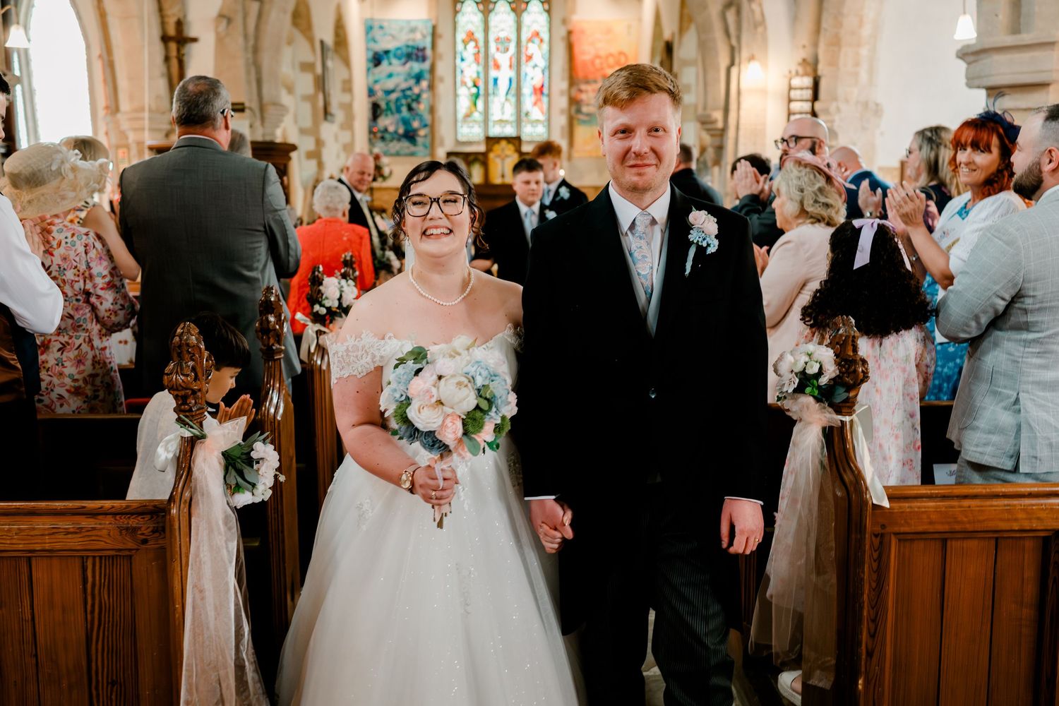 Newlywed couple walks down church aisle smiling, bride in white gown holding bouquet, groom in dark suit, guests celebrating.