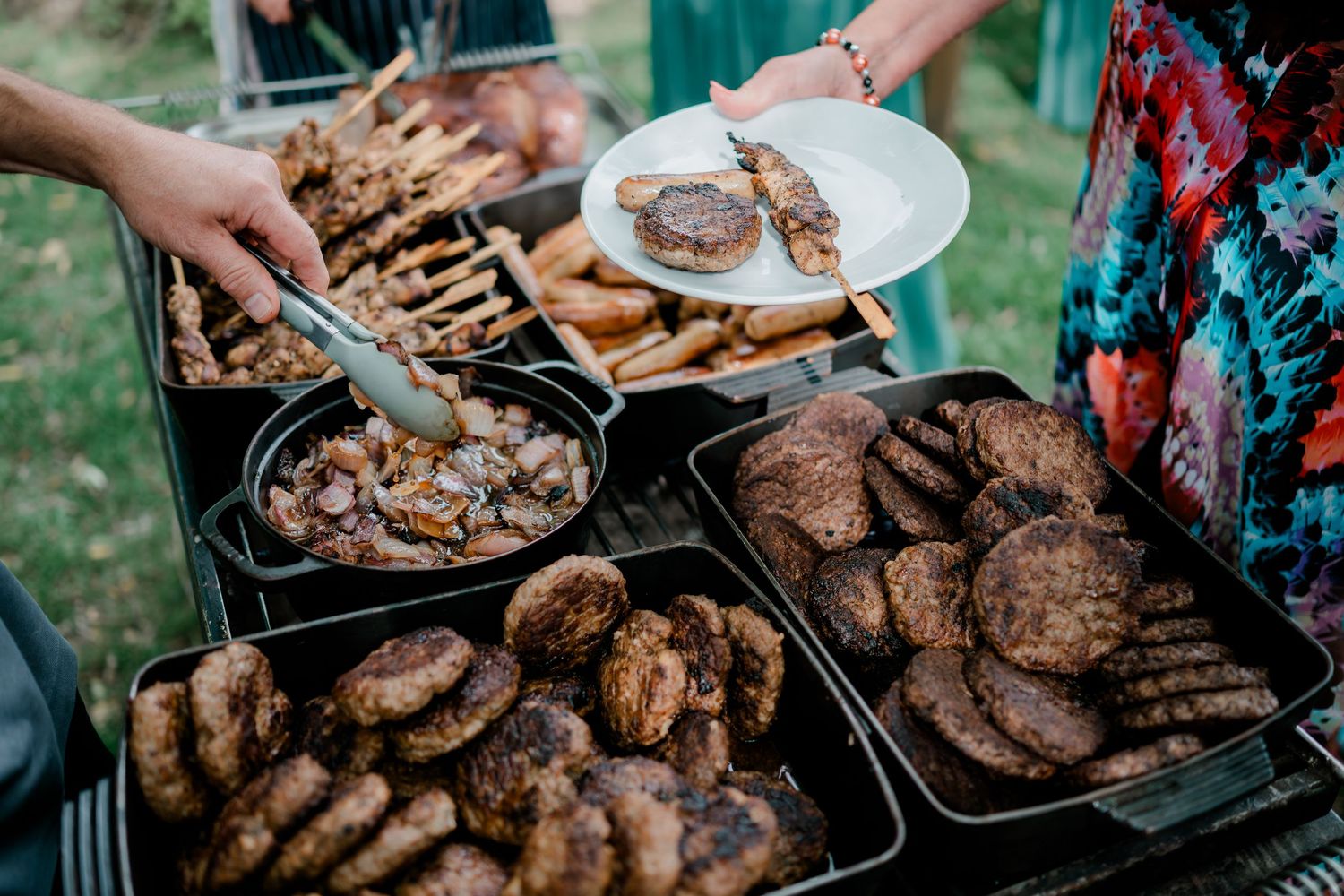 Outdoor BBQ spread with grilled burgers, chicken, and meats on trays, with guests serving themselves using tongs.