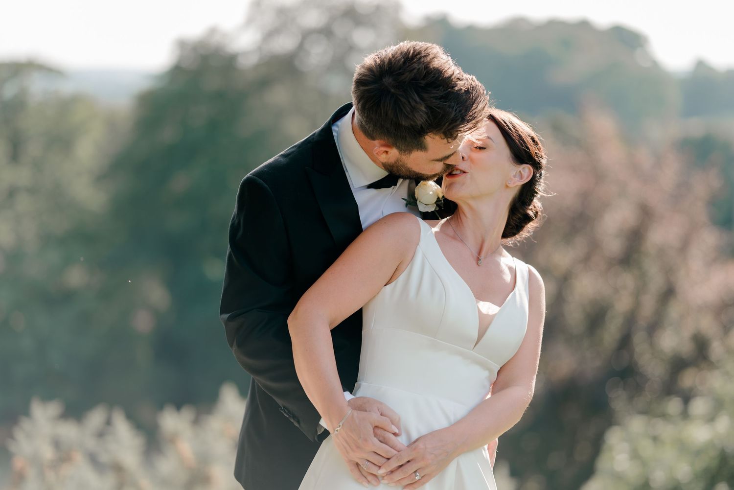 Groom in a dark suit kisses his bride in a white gown as they embrace outdoors with soft green trees blurred in the background.