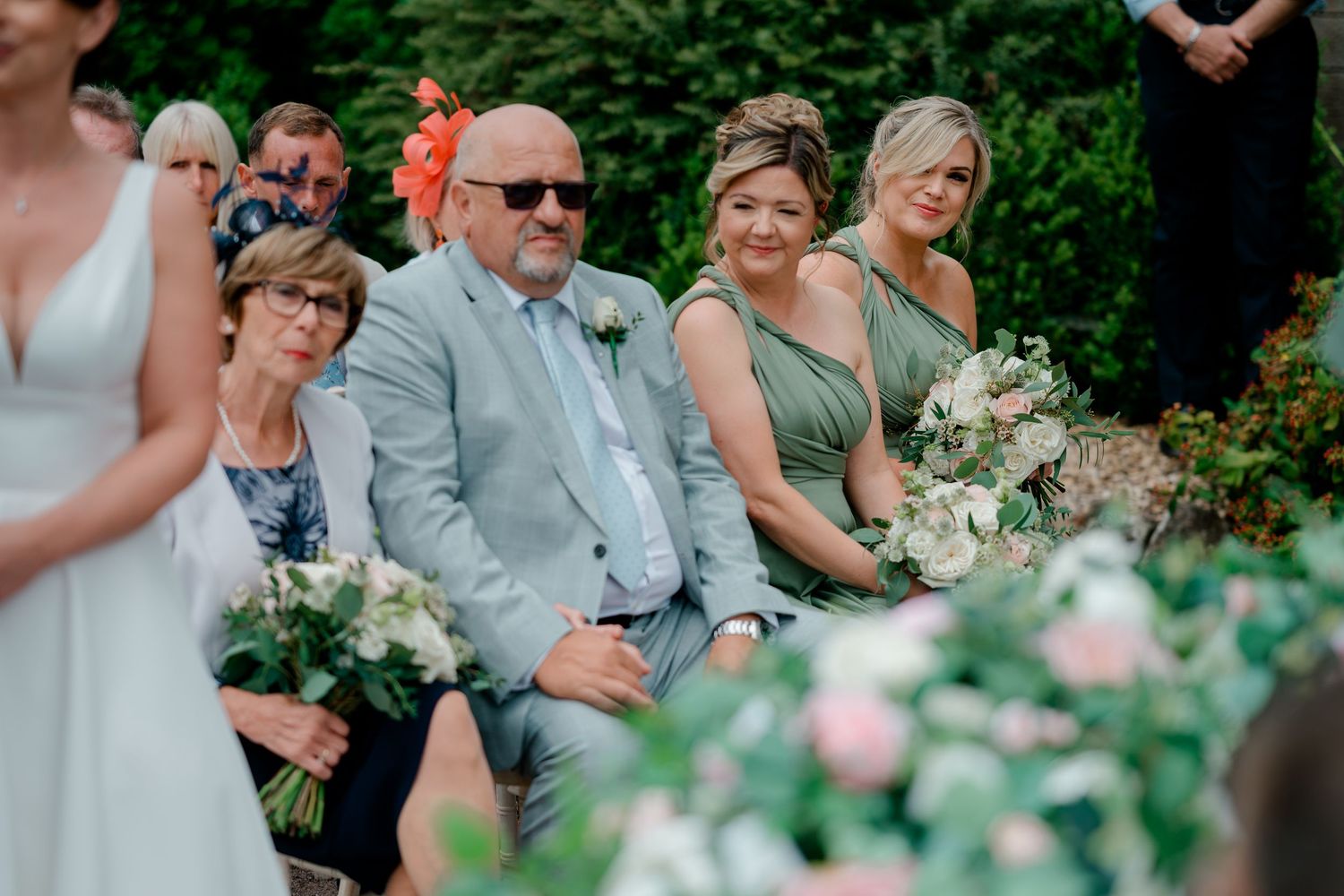Wedding guests in green and gray attire sit smiling among pink and white flower bouquets during an outdoor ceremony.