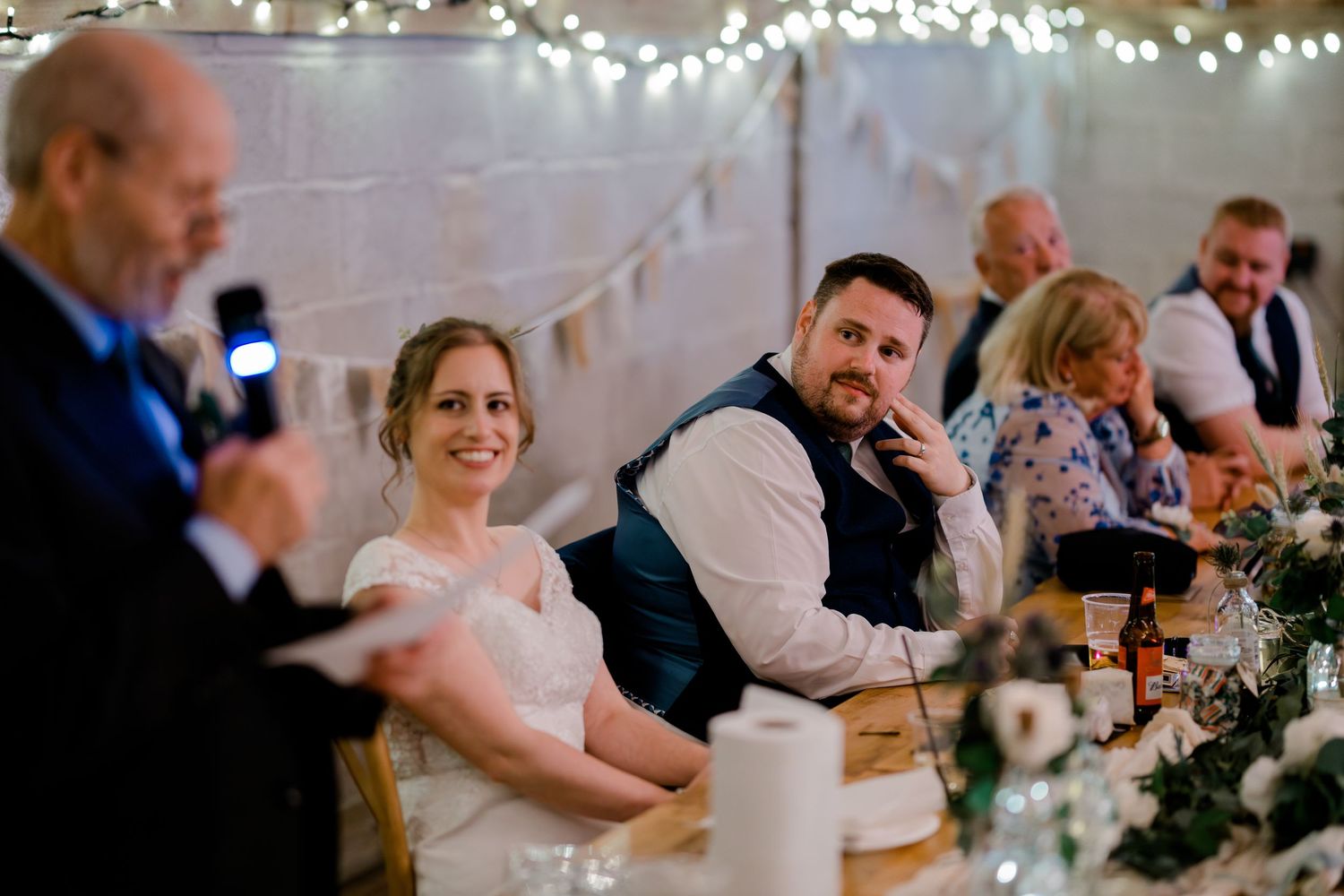 Bride and groom laughing during wedding reception speech in a decorated barn venue with fairy lights.