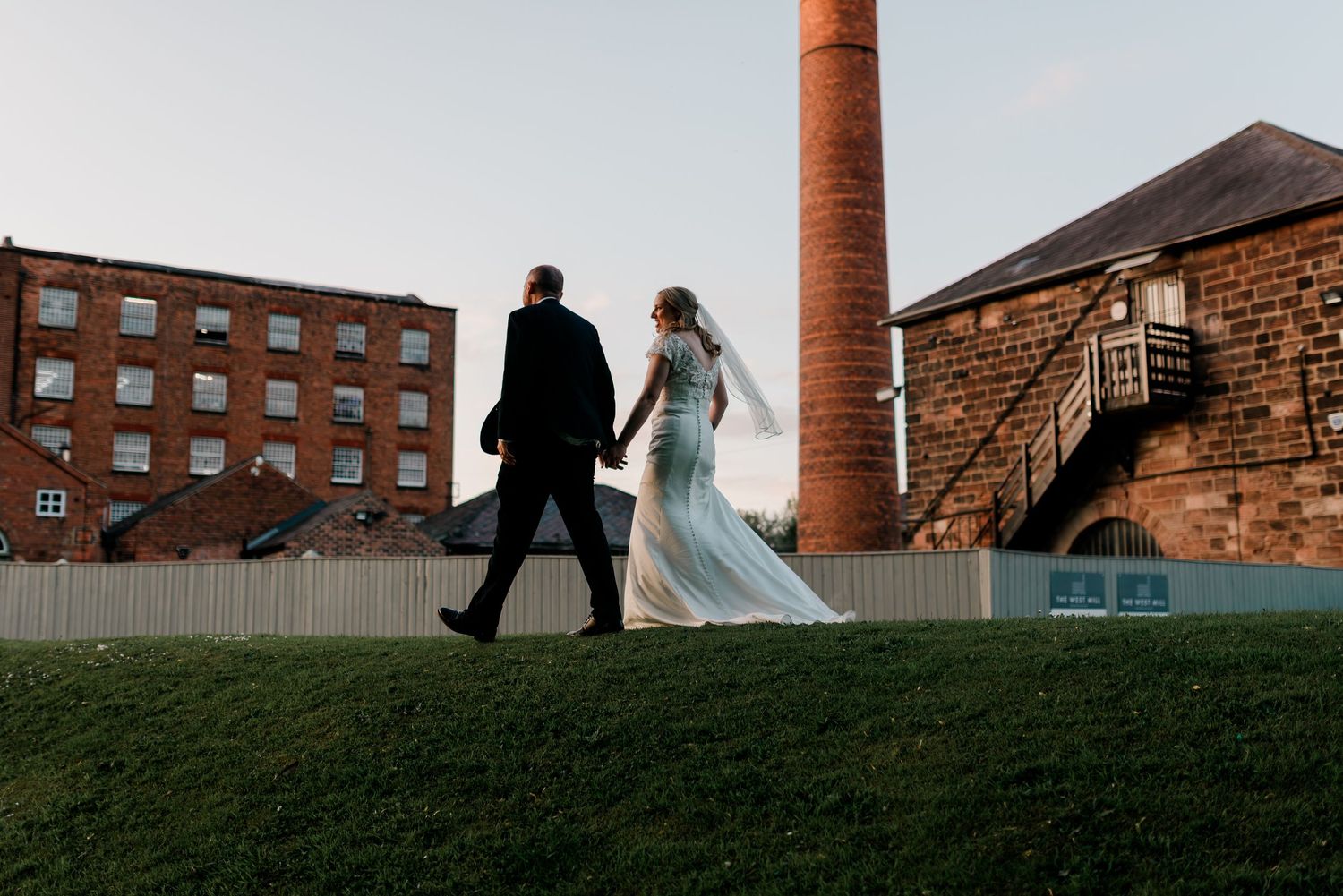 Bride in flowing gown and groom walk hand in hand past historic mill building at dusk.
