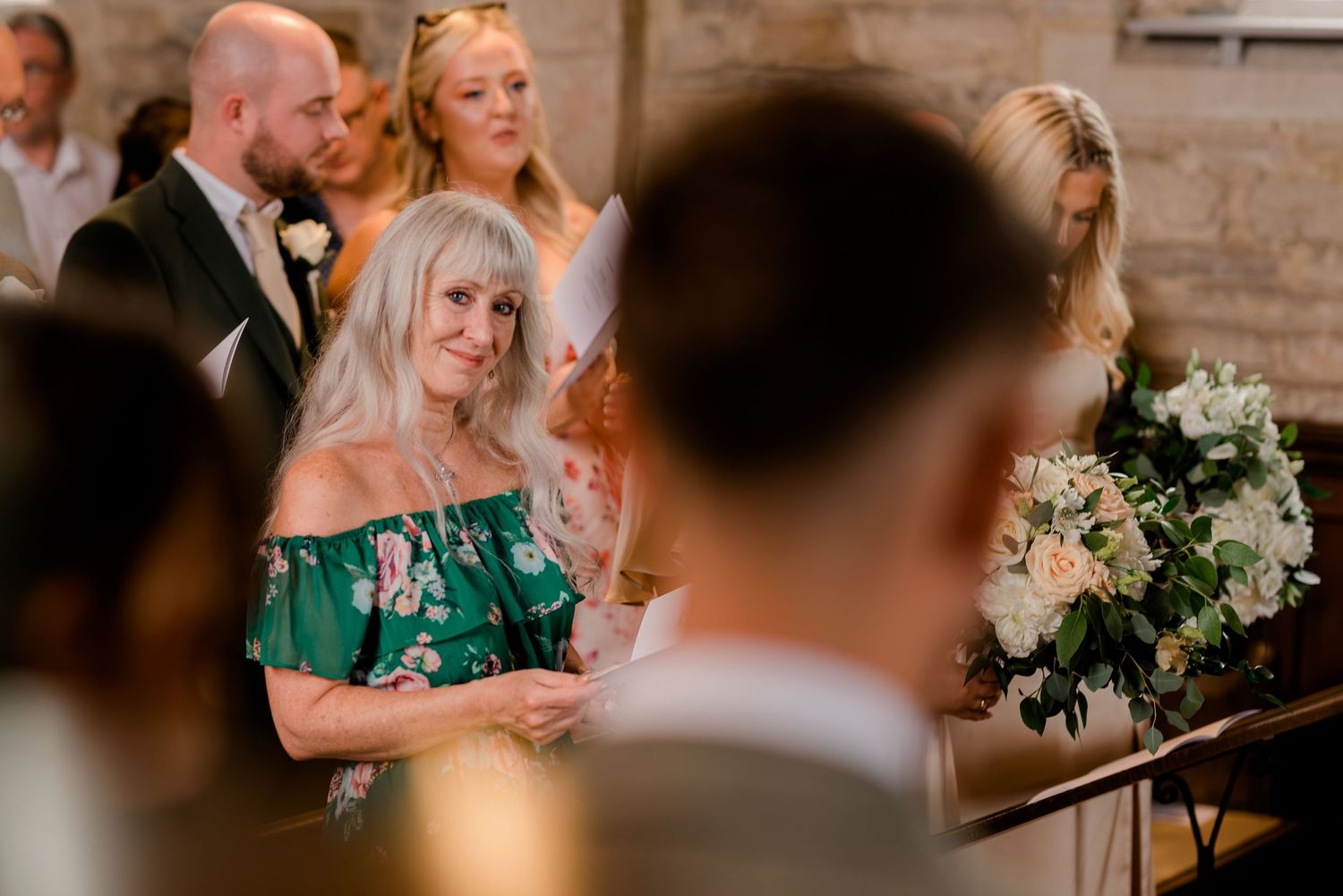 Smiling older woman with blonde hair watching a wedding ceremony with joy inside a stone venue.