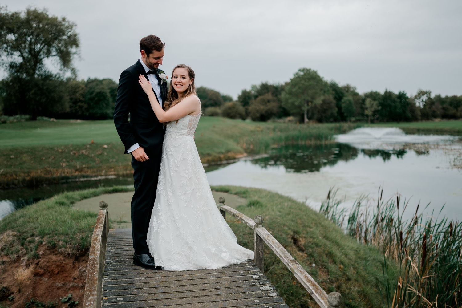 Bride and groom share a tender embrace on a wooden bridge overlooking a serene pond.