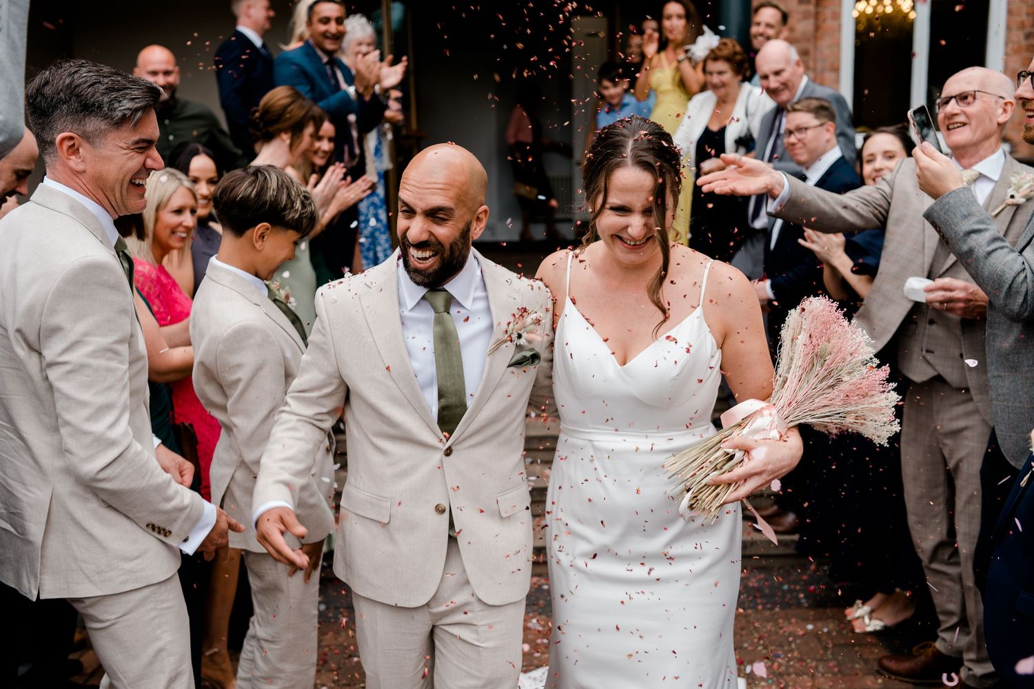 Newlywed couple walks through confetti shower surrounded by cheering wedding guests outdoors.