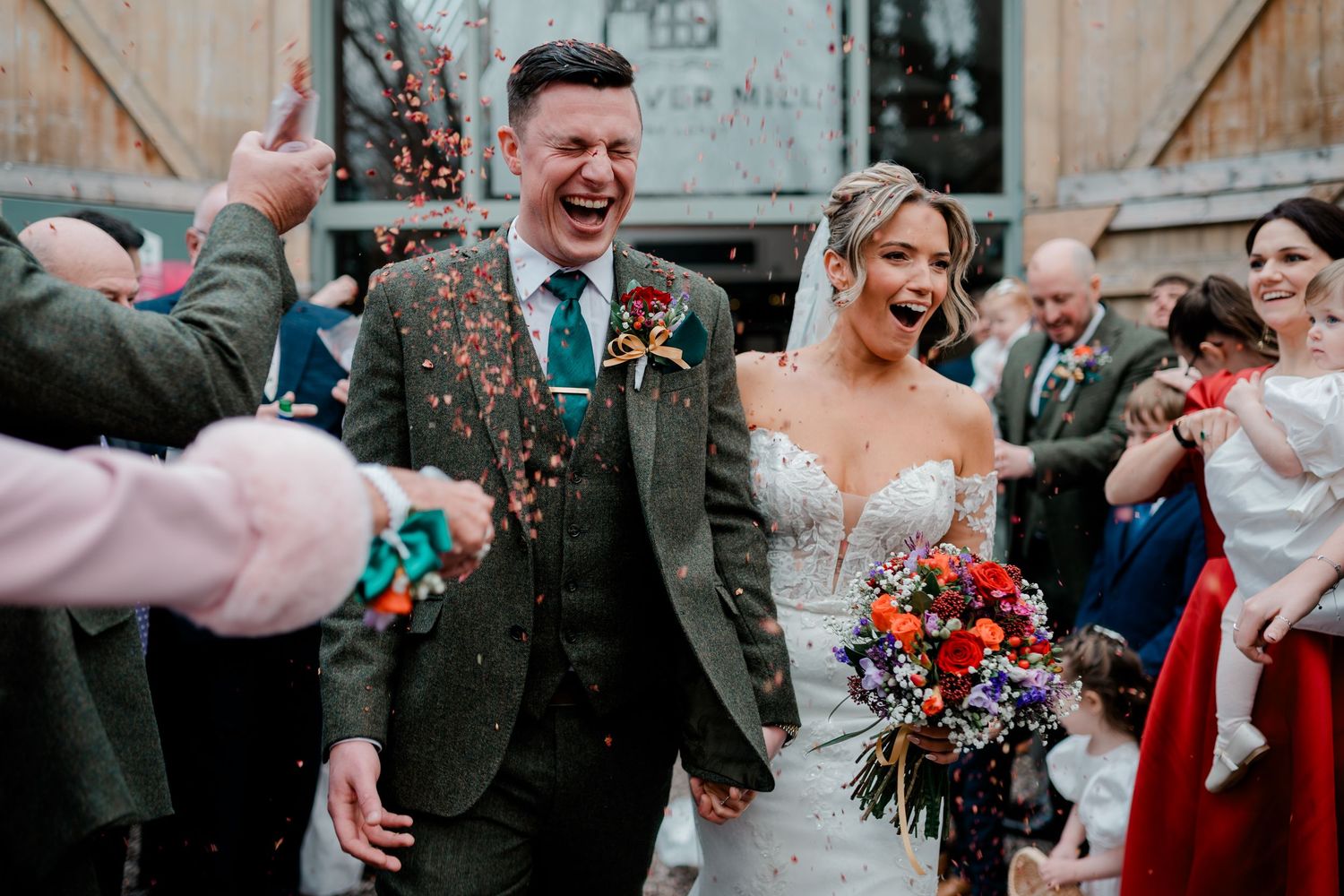 Joyful newlyweds laughing as colorful confetti is thrown outside rustic wedding venue.