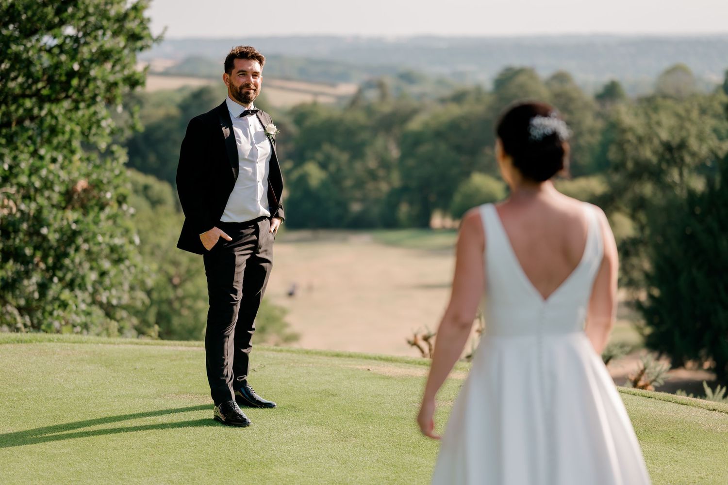 A groom in a black suit and bow tie stands on a grassy hilltop as his bride in a white gown faces him, with rolling countryside behind.