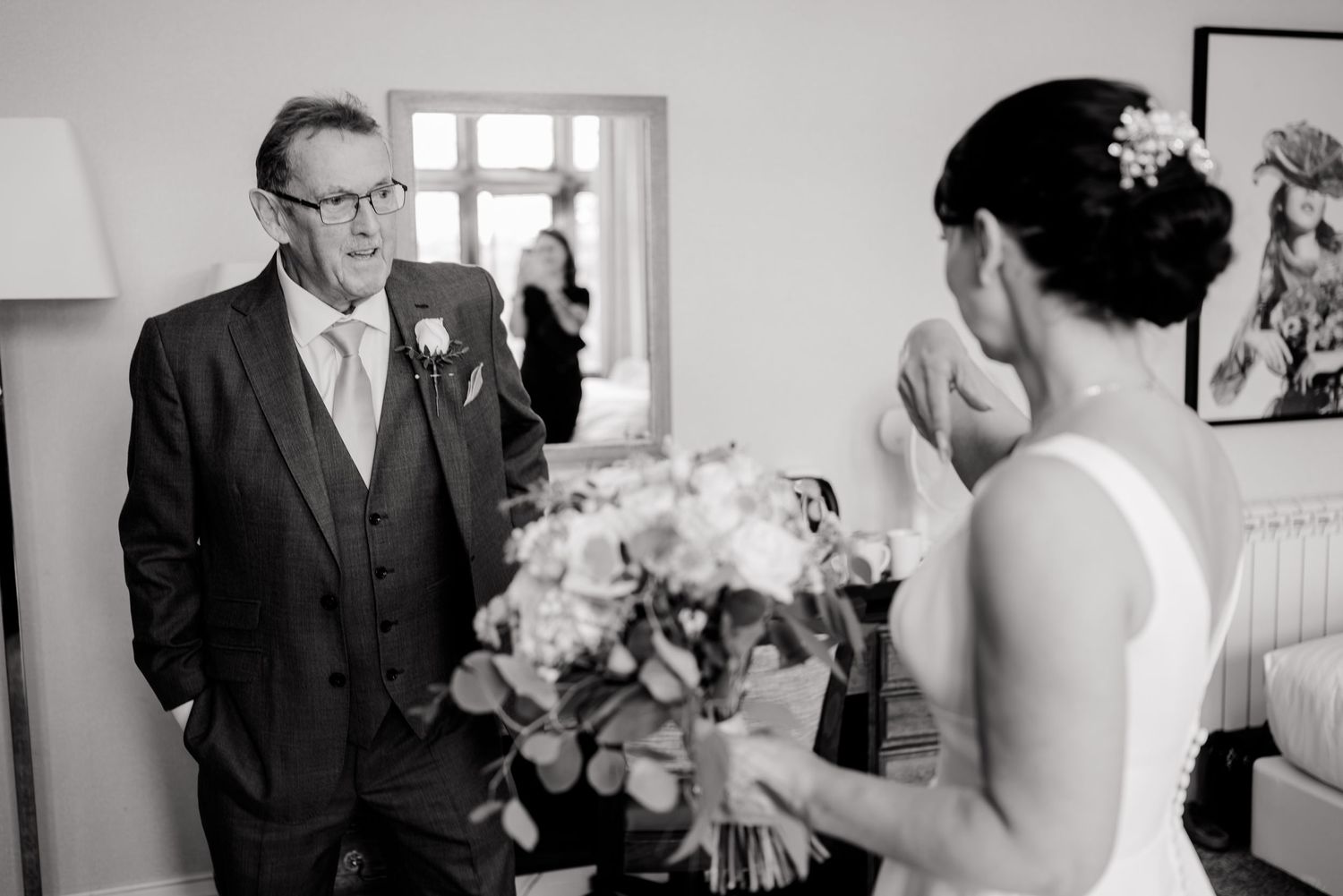 A father in a three-piece suit with a boutonniere looks emotionally at his daughter, a bride holding a bouquet, before the wedding.
