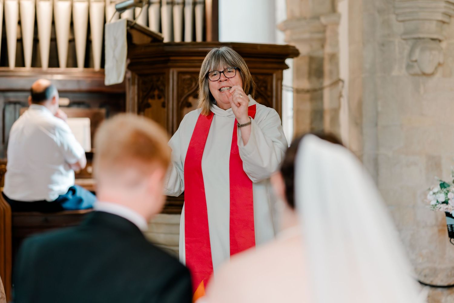 Female officiant in red stole smiling during wedding ceremony inside a historic stone church.
