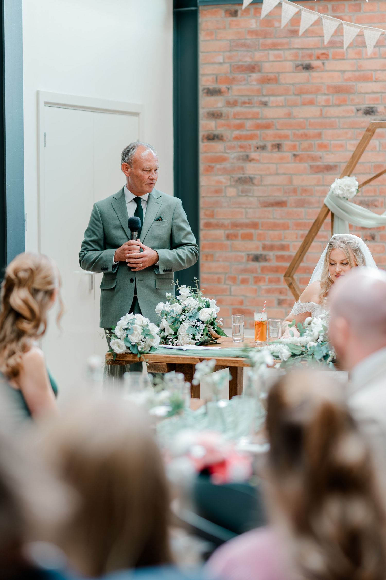 Man in gray suit giving a wedding speech while bride sits at decorated reception table with white floral centerpieces.