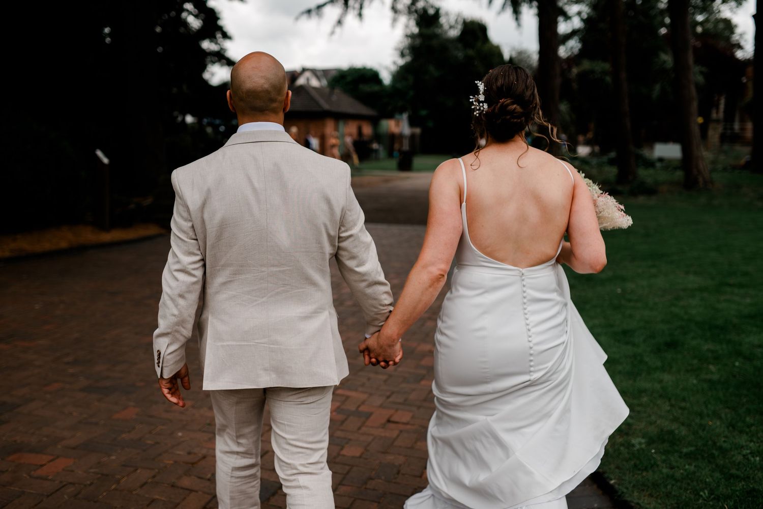 A couple in wedding attire holds hands while walking on an outdoor path surrounded by trees.