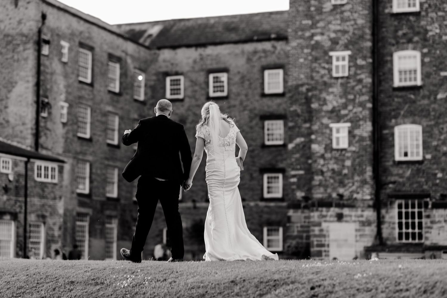 Wedding couple walking along historic stone building facade with arched windows.