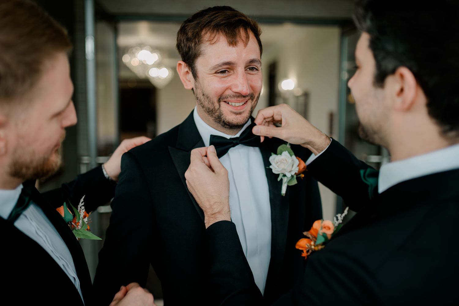 Groomsmen help adjust bow tie and boutonniere before wedding ceremony in indoor setting.