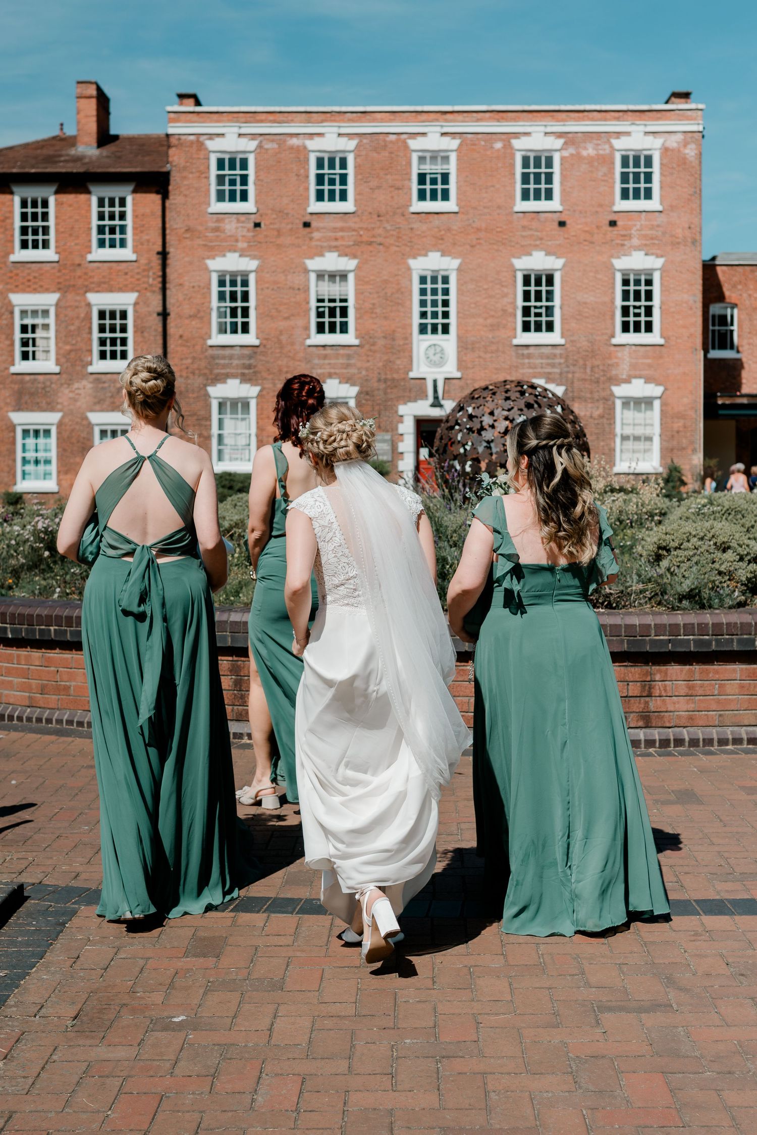 Bride in white gown walks with three bridesmaids in green dresses toward a red brick building on a sunny day.
