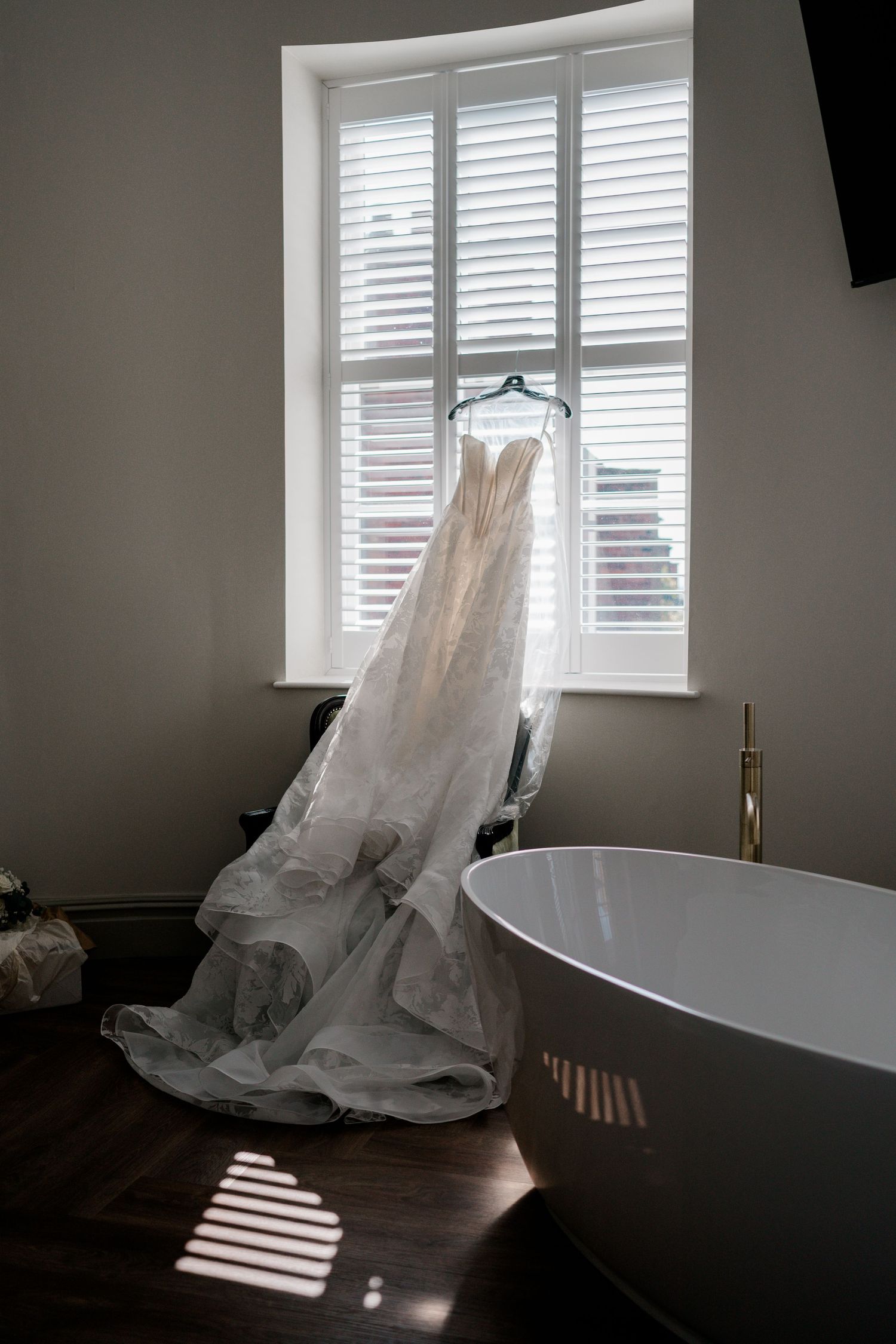 Elegant strapless wedding dress hanging in a sunlit bathroom with a freestanding tub and plantation shutters.