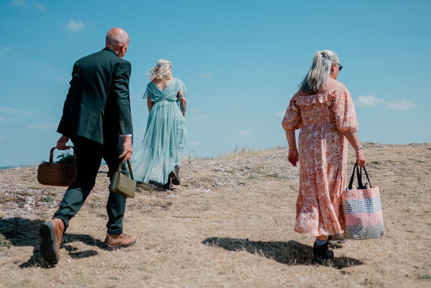 Groom with picnic basket, bride in teal dress, and guest walk across hilltop.