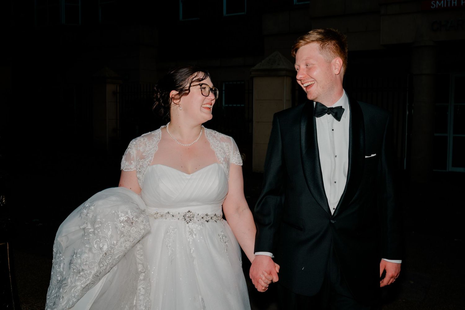 Newlywed couple holding hands and smiling at each other outside at night, bride in lace gown and groom in tuxedo.