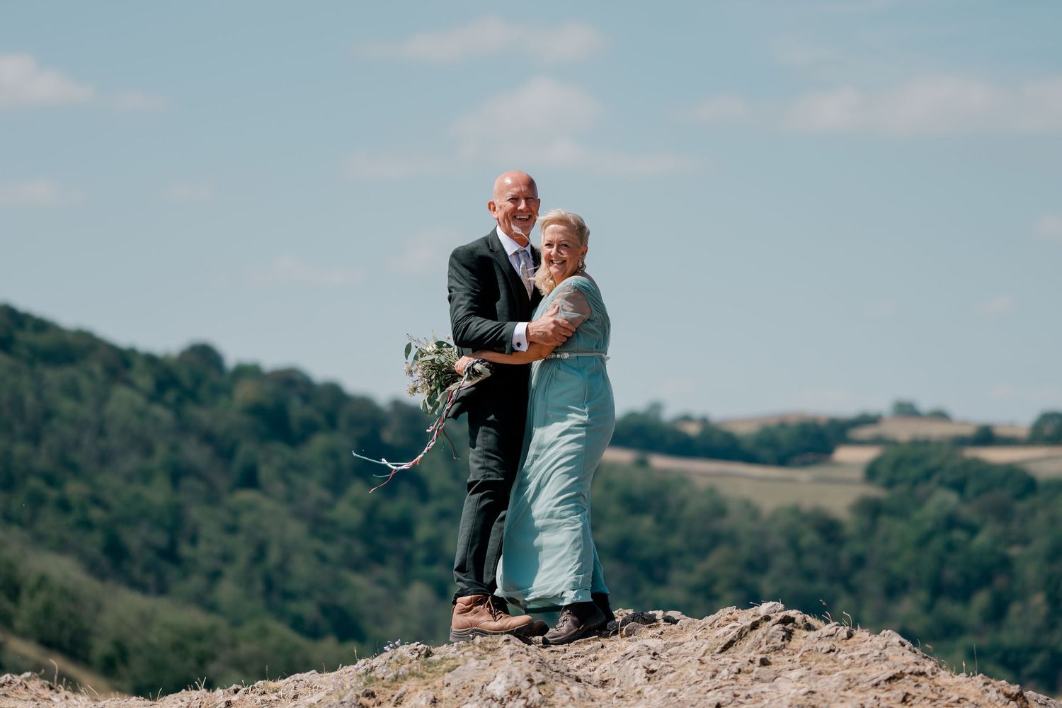 Happy couple embracing on rocky hilltop, groom in dark suit, bride in teal dress, green hills behind.