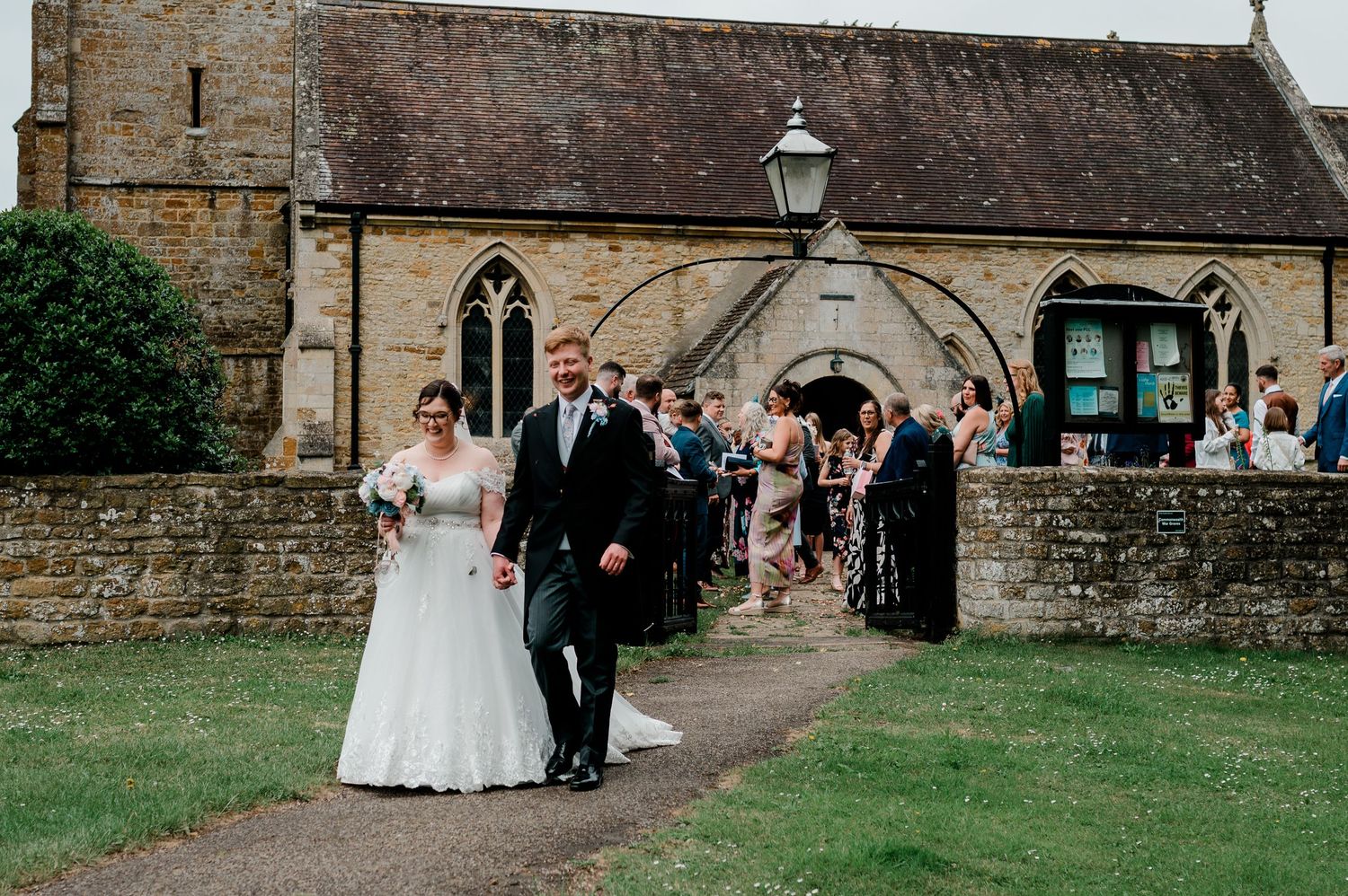 Bride and groom walk out of a stone church surrounded by wedding guests on a sunny day.