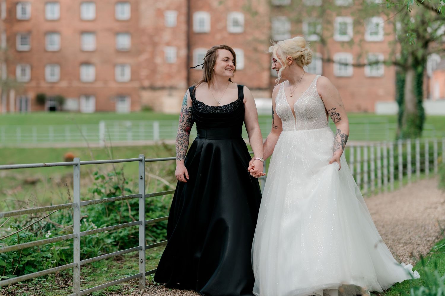 Two brides holding hands, one in black gown and one in white dress, walking outdoors at their wedding.