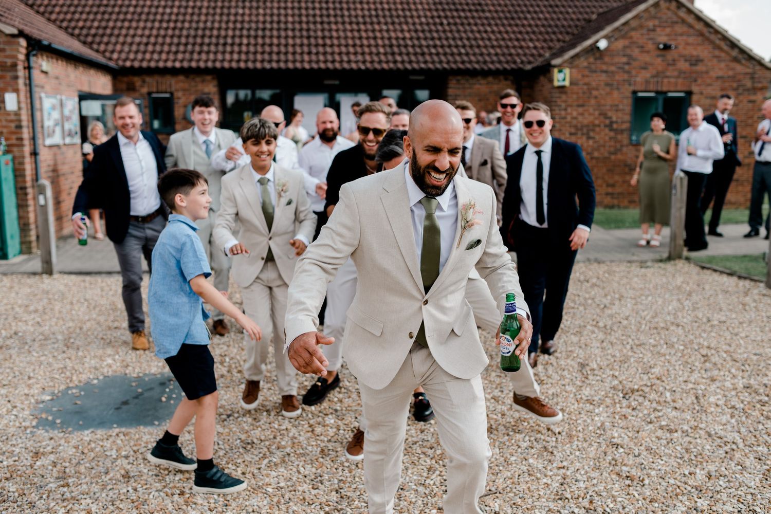 Wedding party walks along a gravel path outside a brick venue building during a celebration.