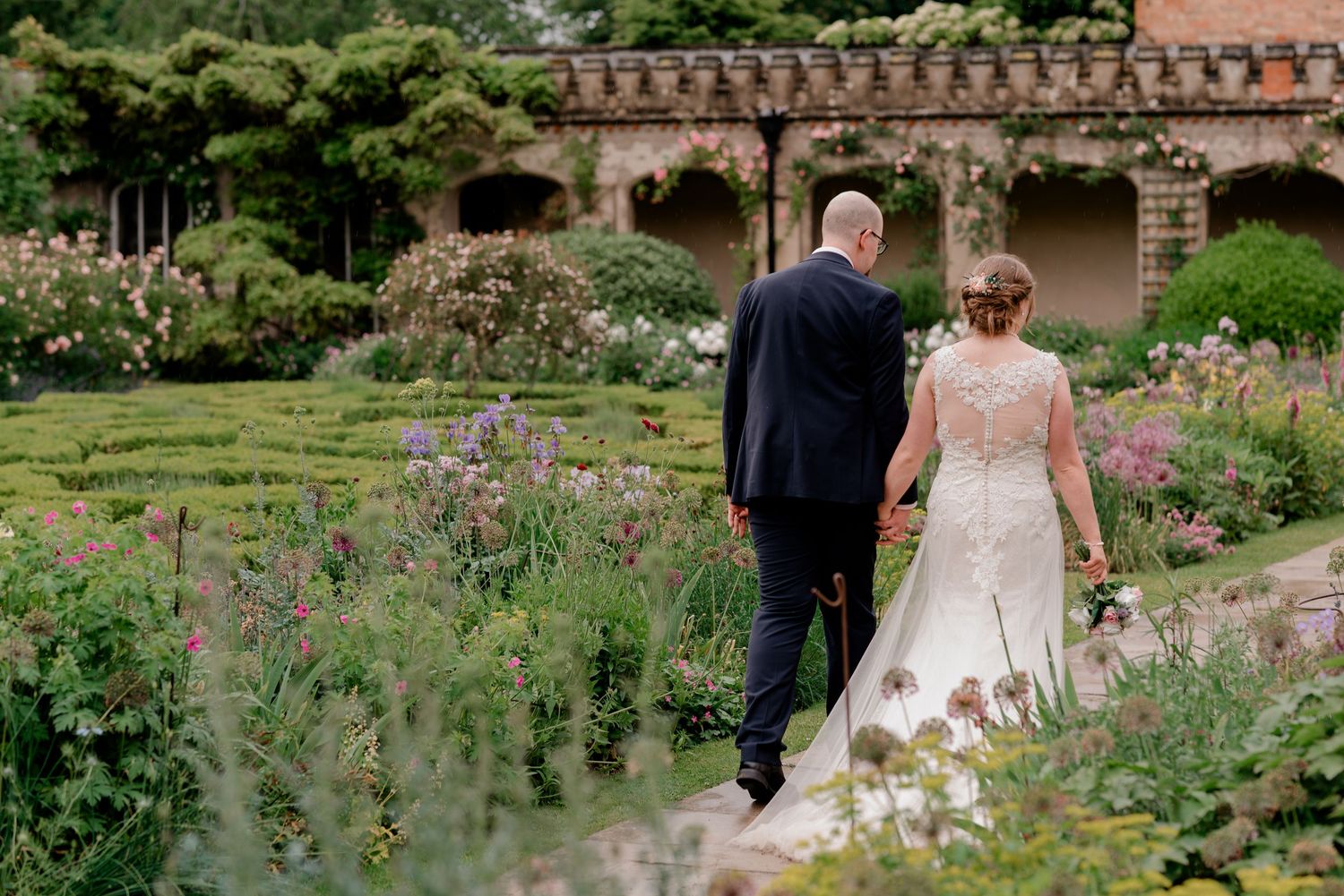 A couple walks through a lush garden path beside a historic brick building with ornate architecture.
