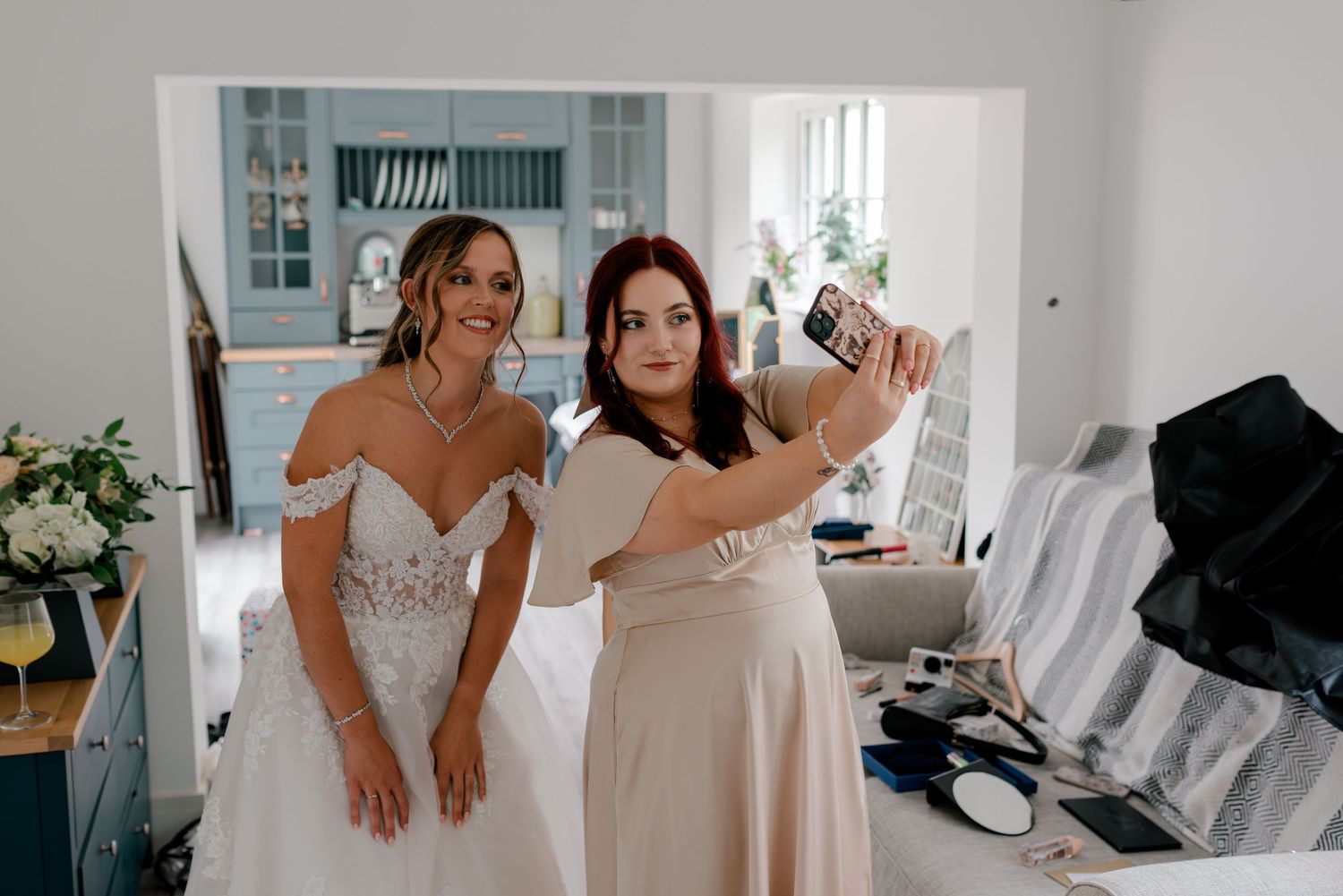 Two people taking a selfie together in a bright room with a wedding dress and formal attire.