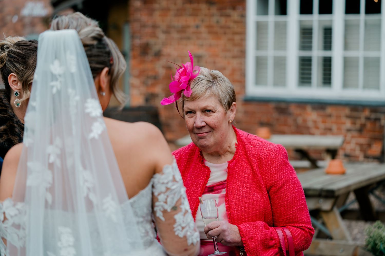 Wedding guest in pink fascinator and red jacket chats with bride during outdoor reception.