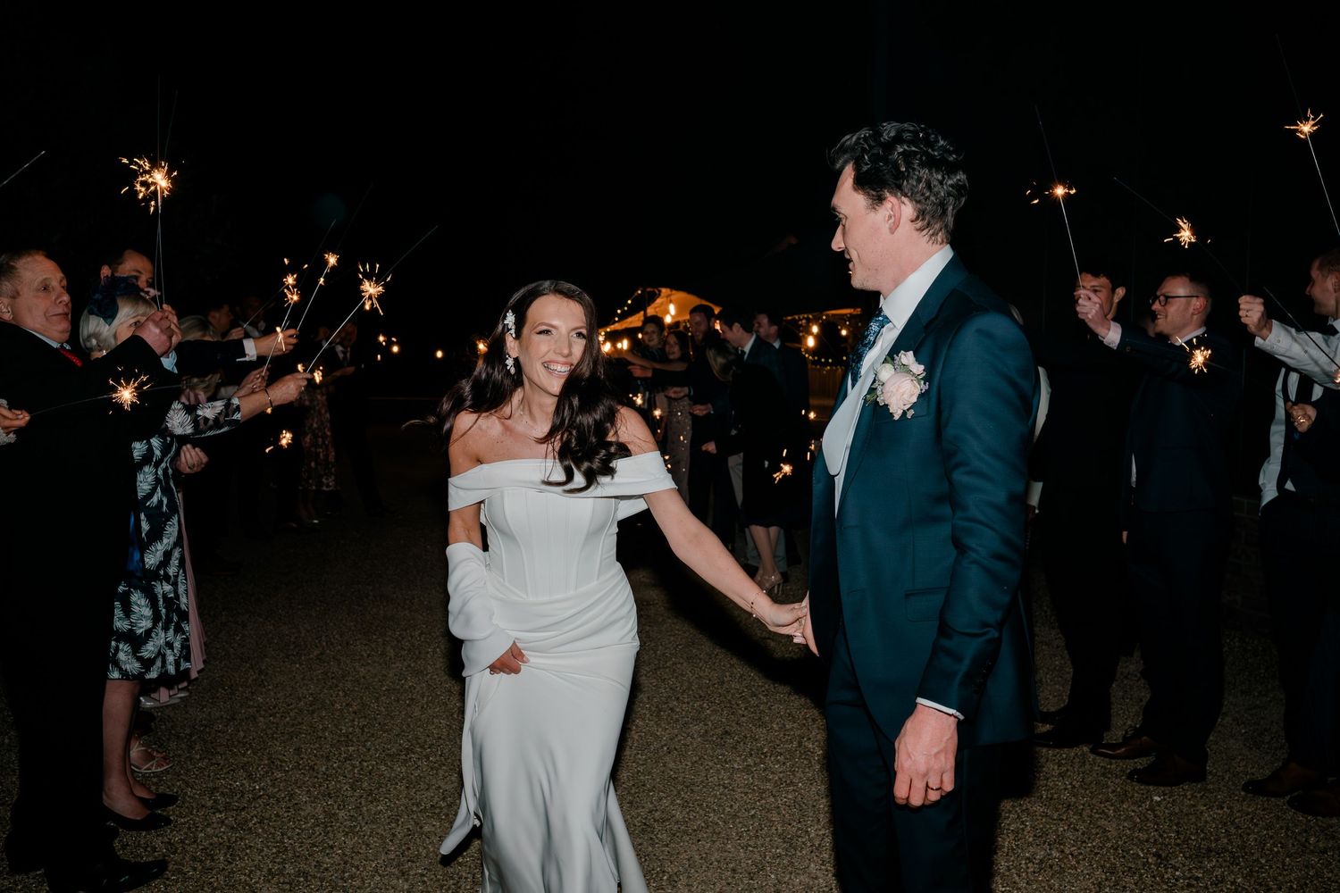 Bride and groom celebrate with sparklers at night surrounded by cheering wedding guests.