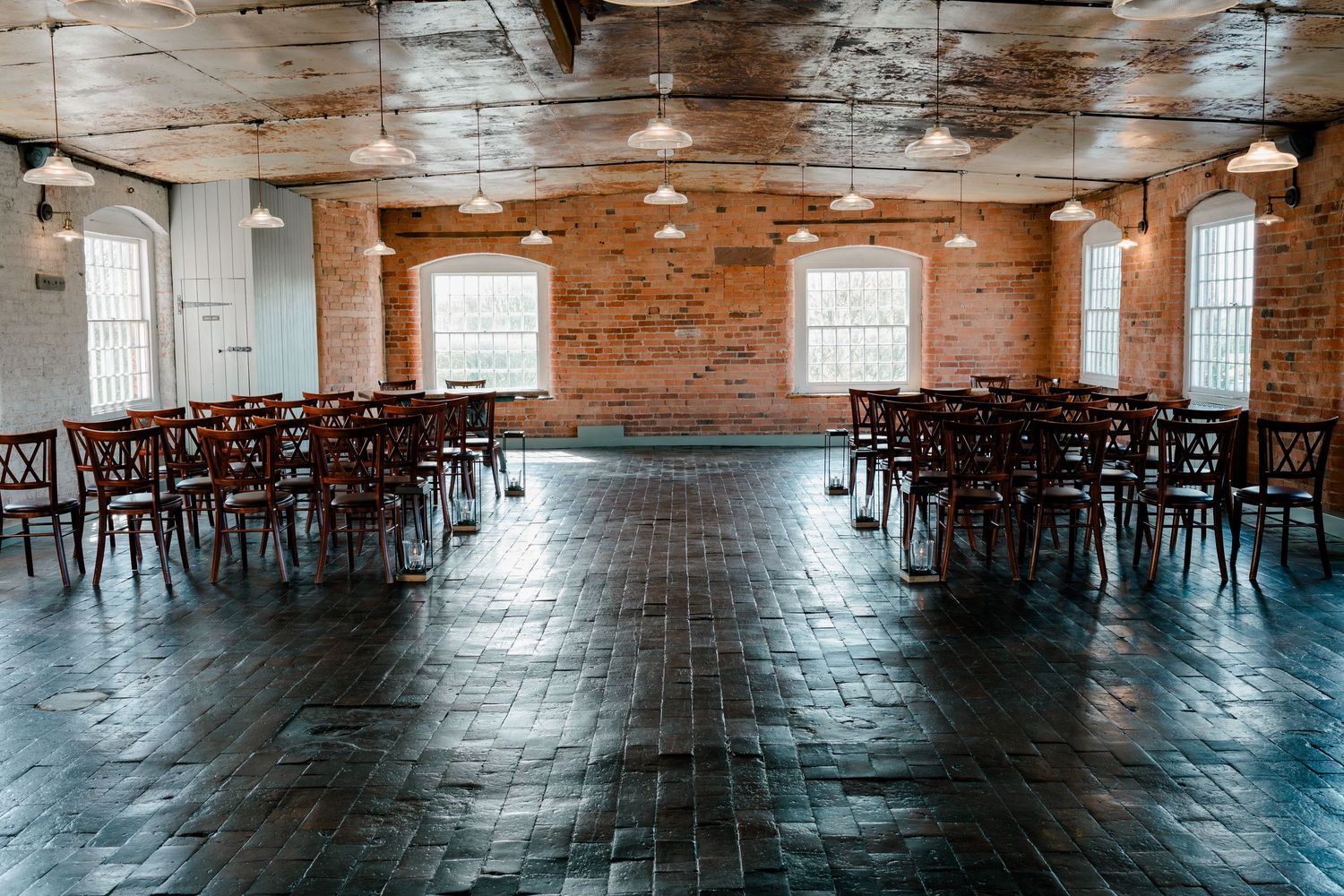 Rustic brick-walled event venue with wooden chairs arranged in rows on dark tile floors, bathed in natural light.