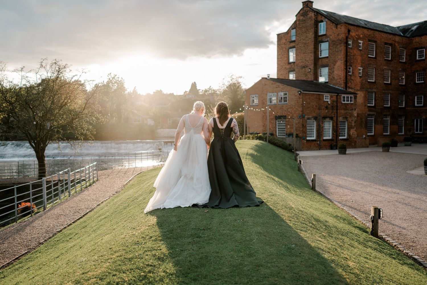 Bride and bridesmaid in black gown walk arm-in-arm on green lawn at golden hour beside a historic brick mill.