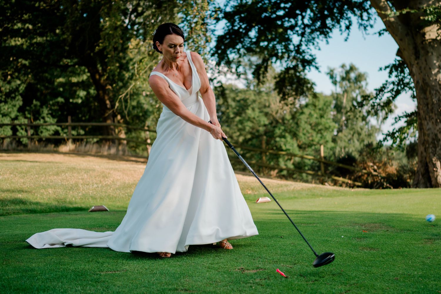 Bride in white wedding gown playing golf on a green course, swinging a club near a large tree.