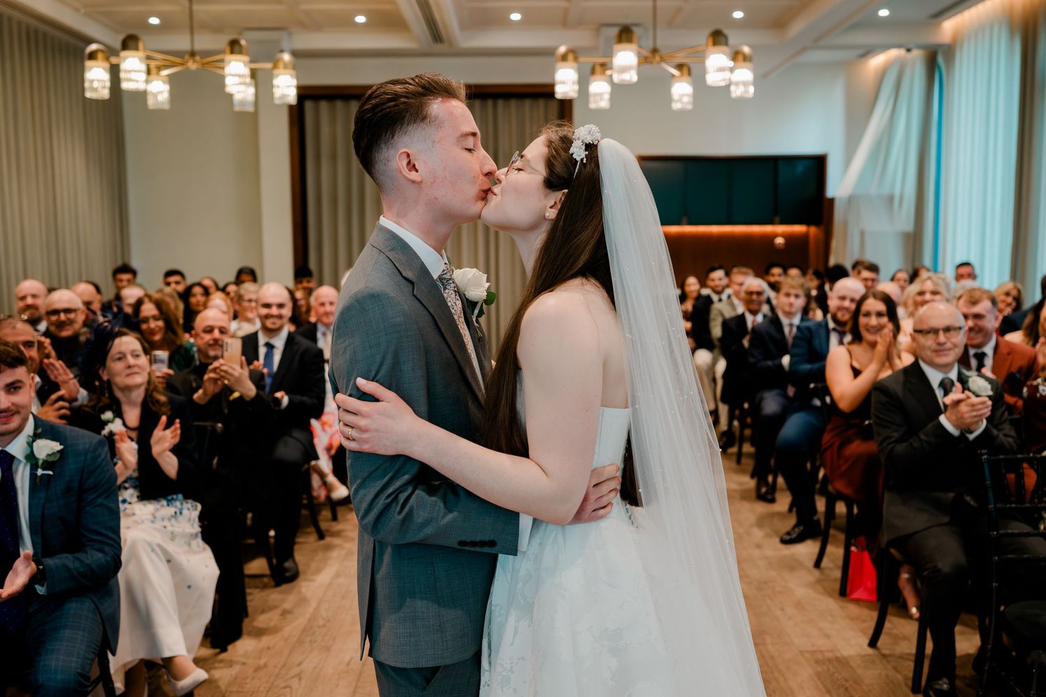Bride and groom share first kiss at wedding ceremony while guests applaud in elegant venue.