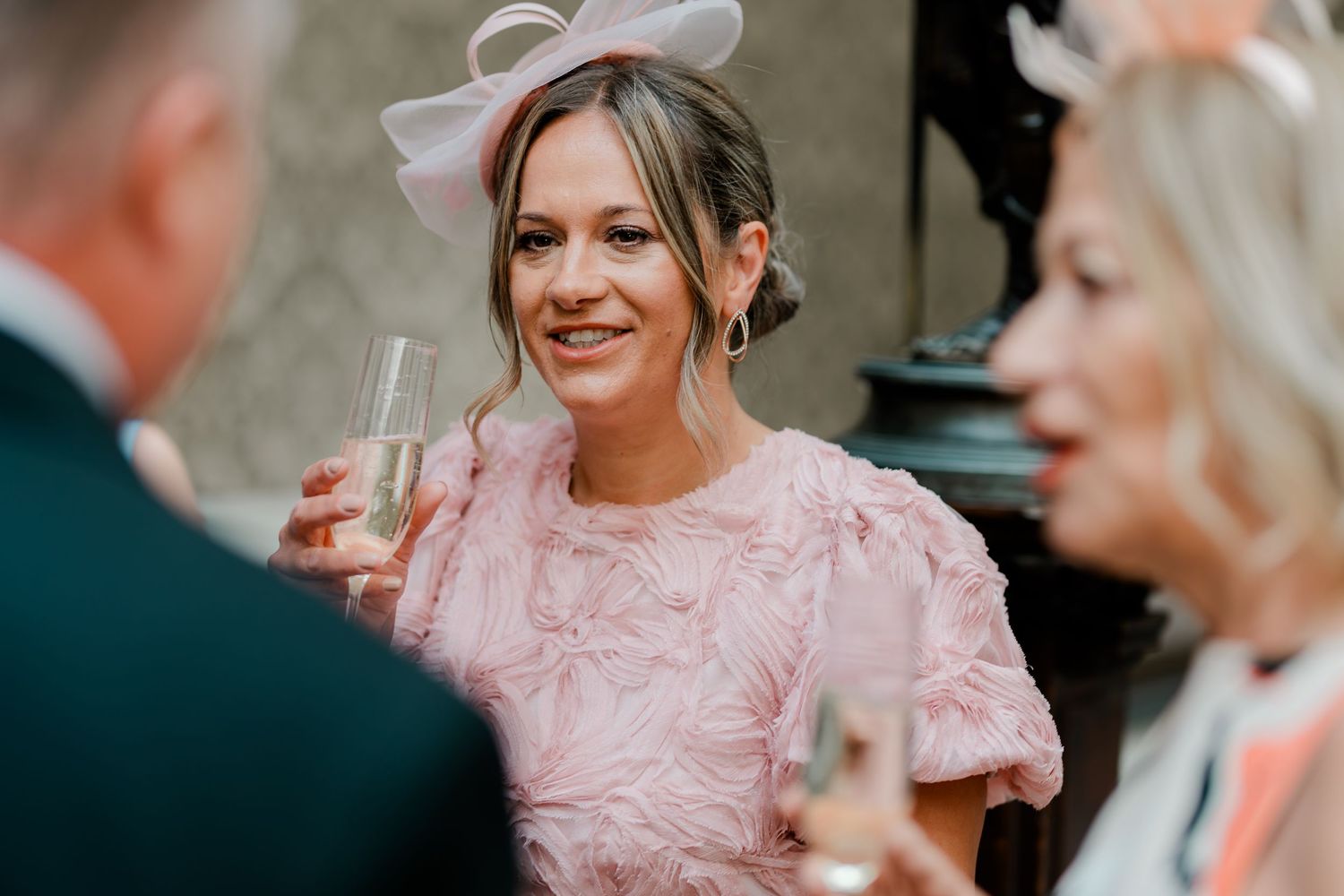 A smiling woman in a pink dress and fascinator hat holds a champagne glass at an elegant wedding reception.