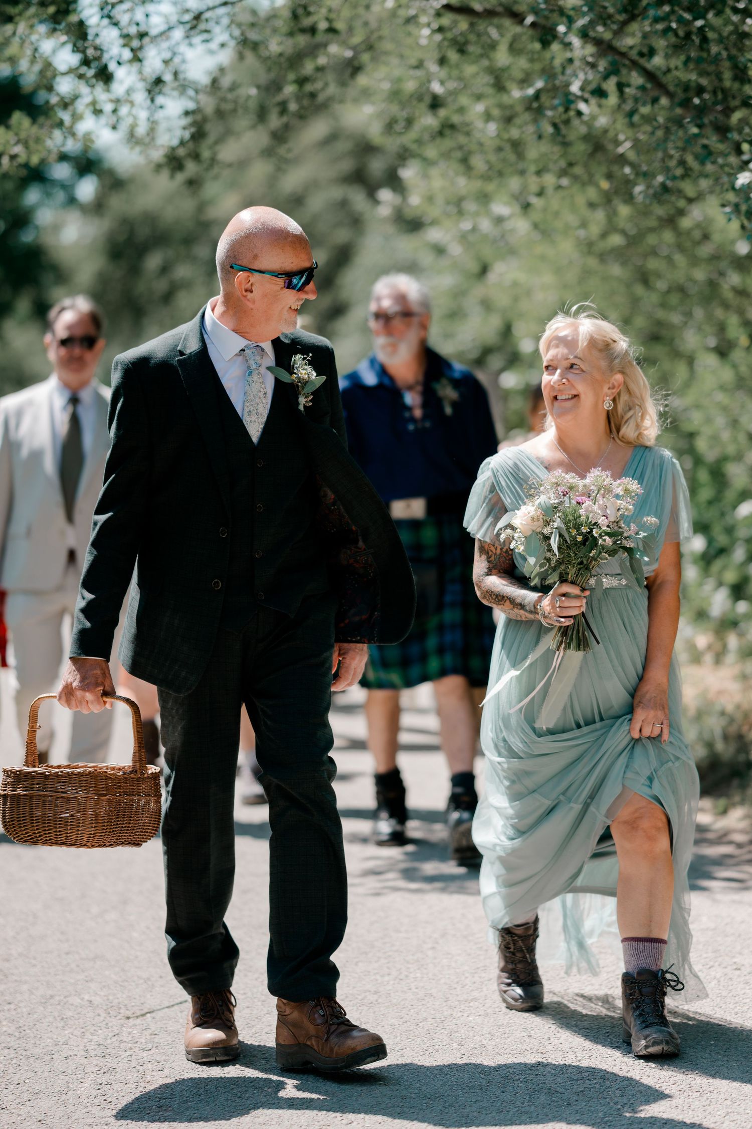Couple walking together at outdoor wedding, man in dark suit holding basket, woman in mint dress with bouquet, guests in background.