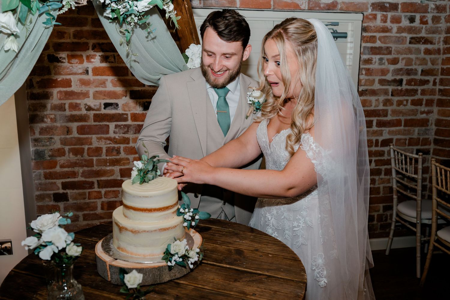 Smiling bride and groom cutting a three-tier wedding cake decorated with white flowers at rustic brick venue.