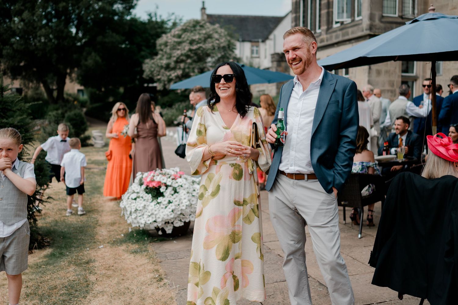 Smiling woman in a floral dress and sunglasses stands beside a man in a blue blazer at an outdoor garden wedding reception with guests.