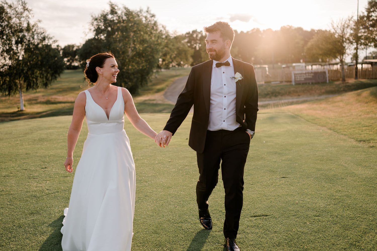 Bride in a white gown and groom in a black tuxedo hold hands while walking across a sunlit grassy field at golden hour.