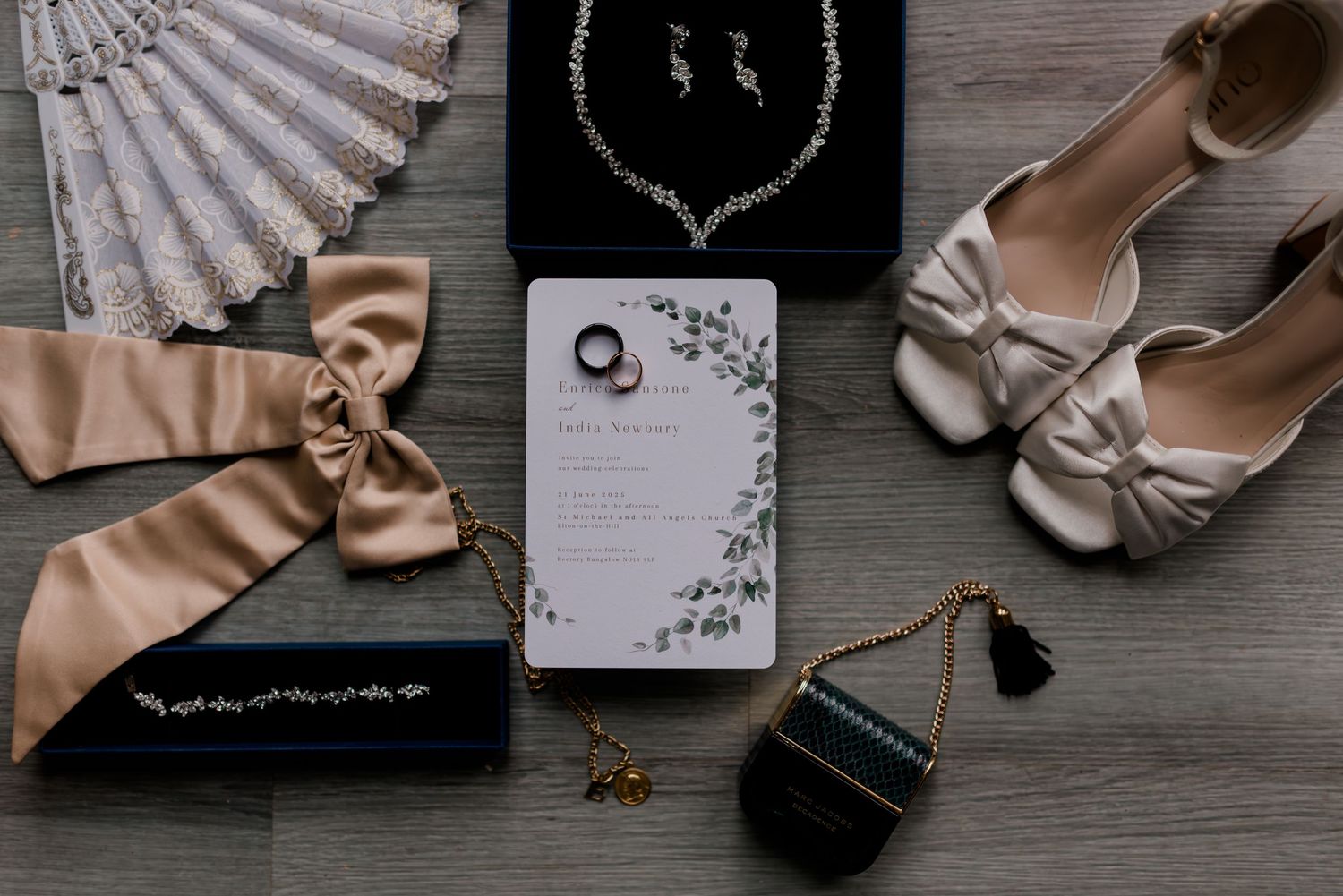 Elegant wedding accessories including fans, jewelry, shoes and invitations arranged on rustic wooden surface.