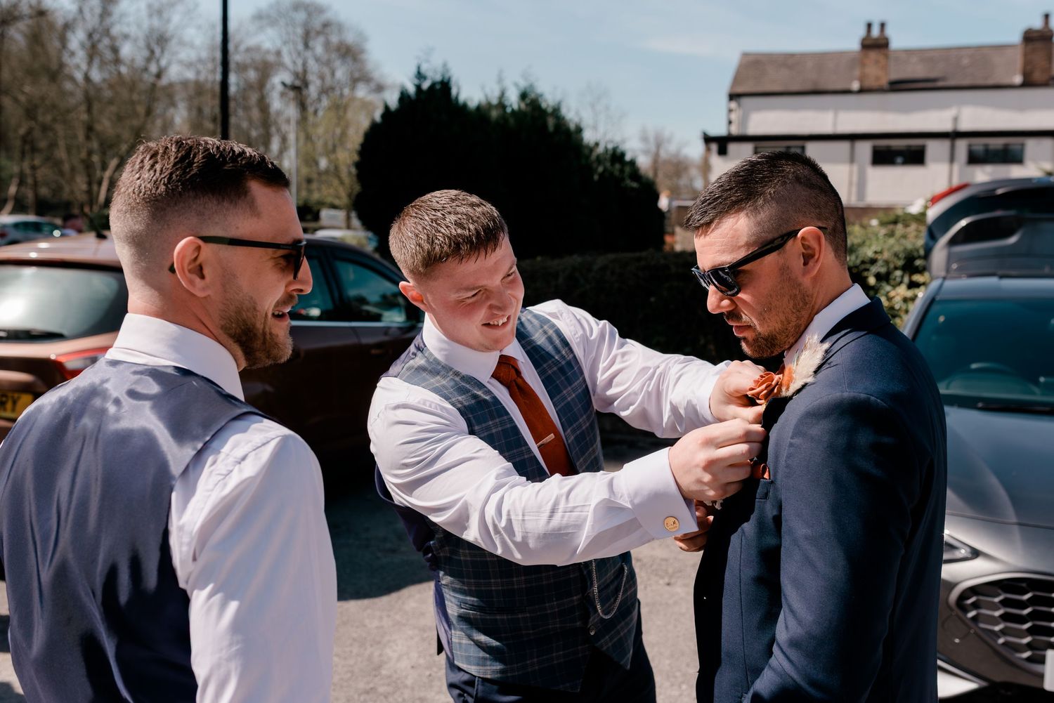 Three groomsmen in formal attire help each other prepare by adjusting ties in an outdoor setting.
