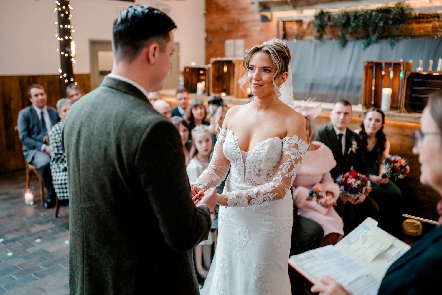 Bride in lace off-shoulder gown exchanging vows with groom in green tweed suit indoors.