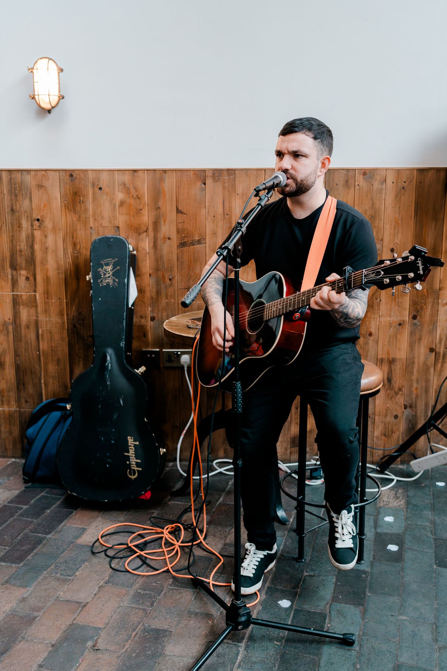 Tattooed acoustic guitarist performs live wedding entertainment at rustic indoor venue.