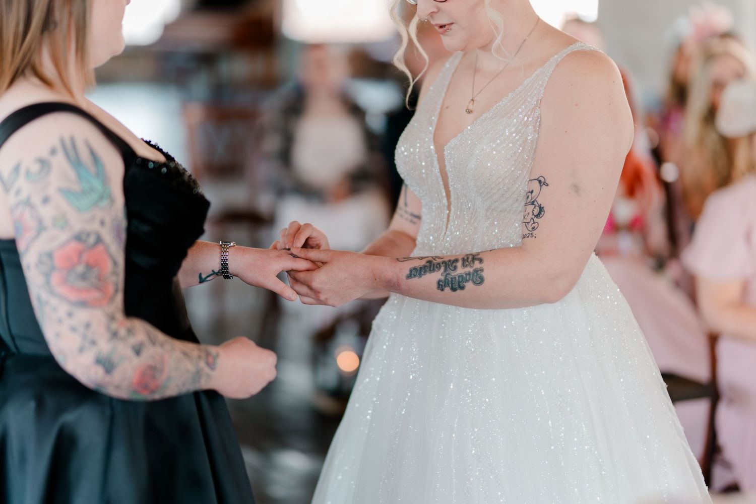 Bride in white gown exchanging rings with tattooed bridesmaid in black dress during wedding ceremony.