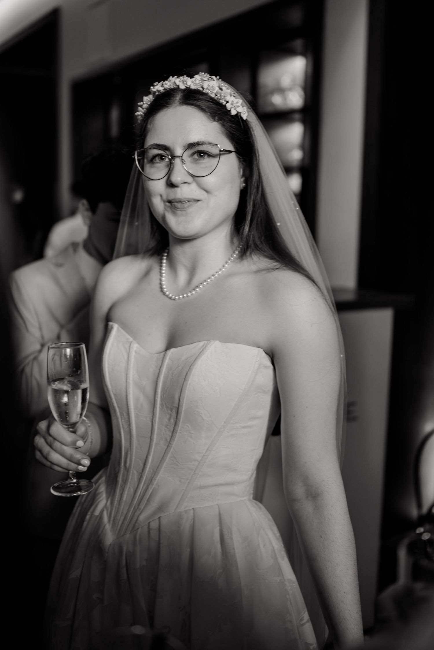 Bride in strapless gown, floral tiara, veil, pearl necklace, and glasses holding champagne glass at wedding reception.